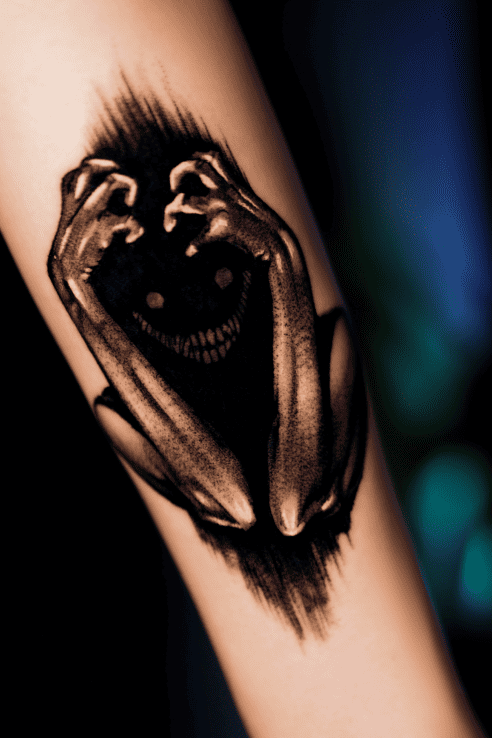 Horror creature covering its grinning face with clawed hands, black and grey forearm tattoo